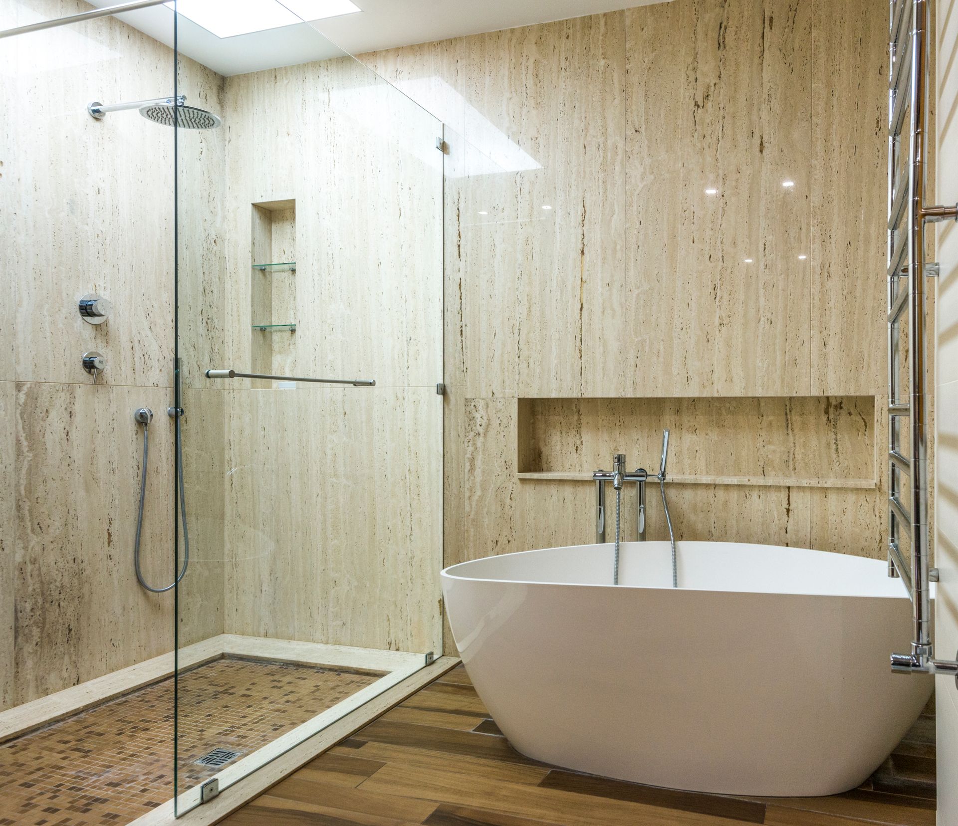 Spa-style primary bathroom in honed beige travertine-look slabs under a skylight, with a frameless glass walk-in shower, a glass mosaic shower floor, a white freestanding tub and a wood-look plank tile floor