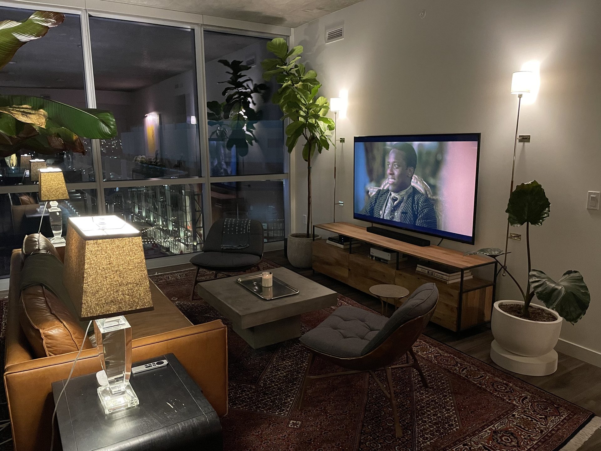 Wall-mounted TV over a low oak media console in a downtown Phoenix condo at night, flanked by floor lamps with the city lights through floor-to-ceiling glass