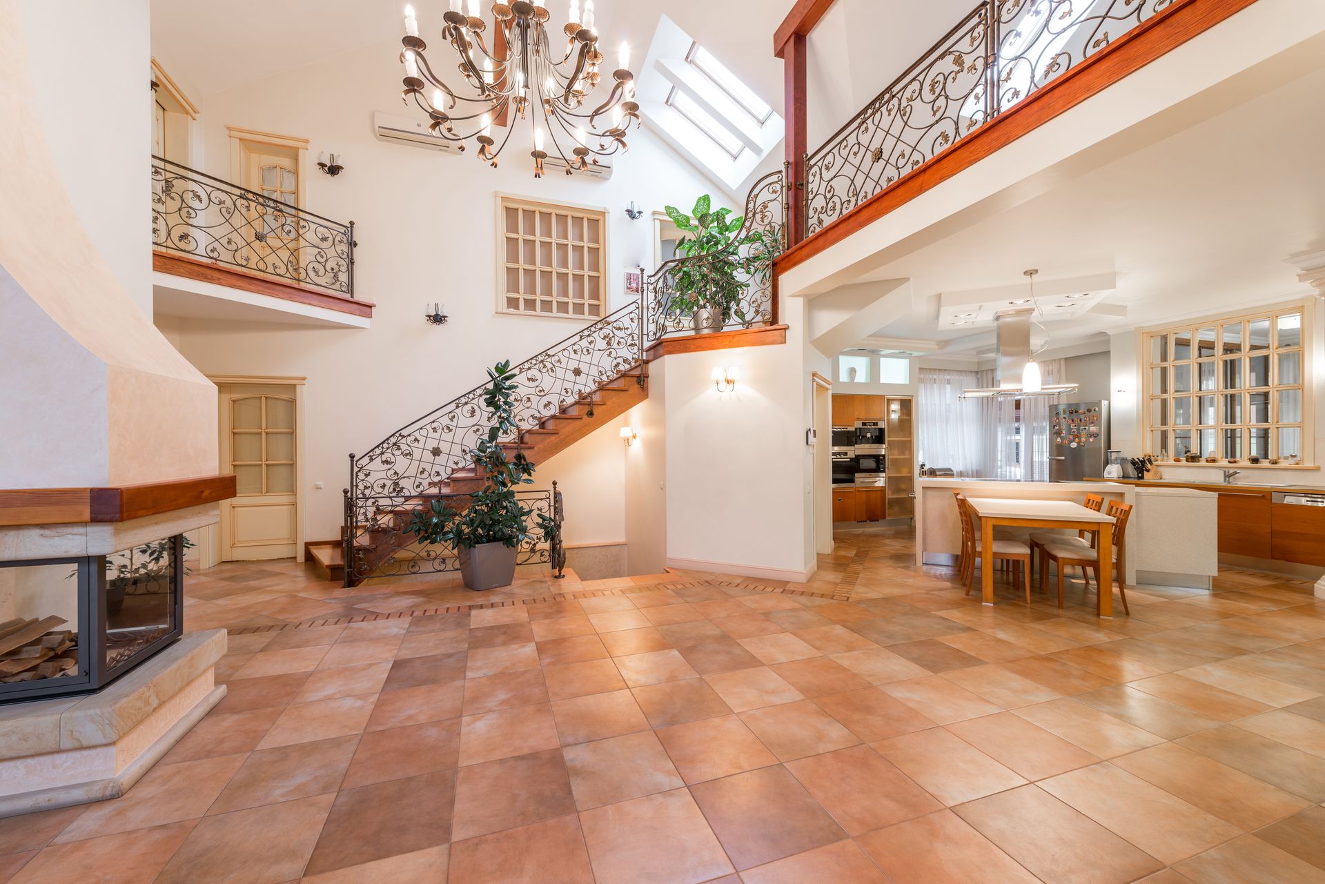 Bronze wrought-iron candle-style chandelier hanging high in a two-story entry hall with a vaulted ceiling, skylights, a curved wood staircase with scrolled iron railings and an upper balcony