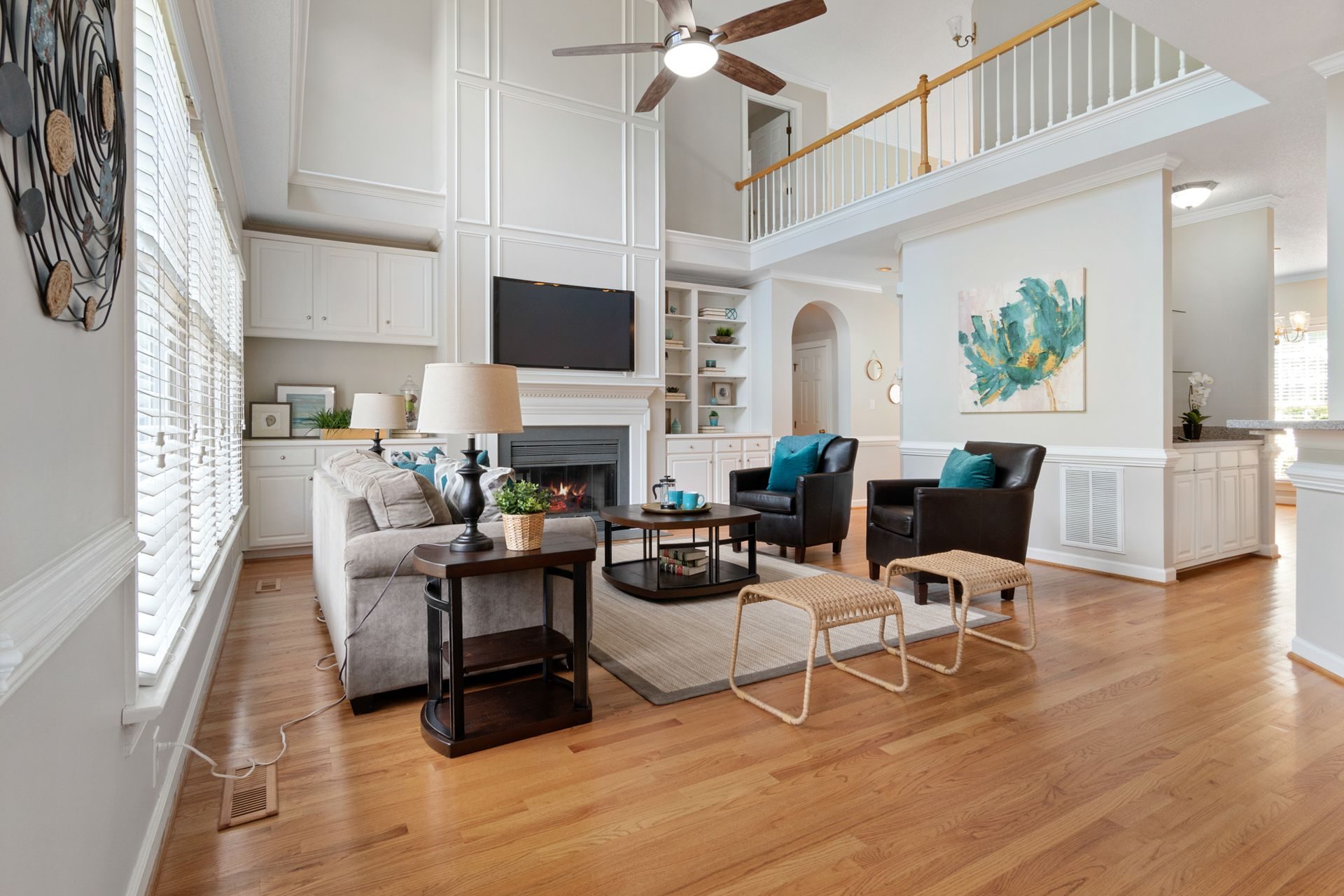 A two-story great room with oak floors, a gray sofa, leather chairs and a fireplace with a TV and built-in shelves, under an open loft balcony railed with white balusters and a stained wood cap