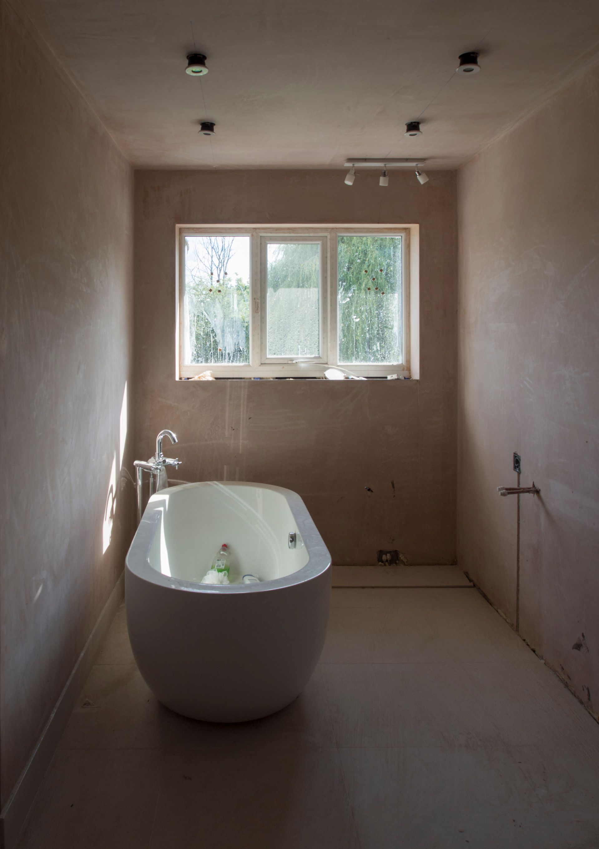 An unfinished bathroom with skim-coated walls not yet painted, a freestanding soaking tub and floor-mount filler set in place, plumbing stubs and an empty electrical box in the wall, and a bare subfloor