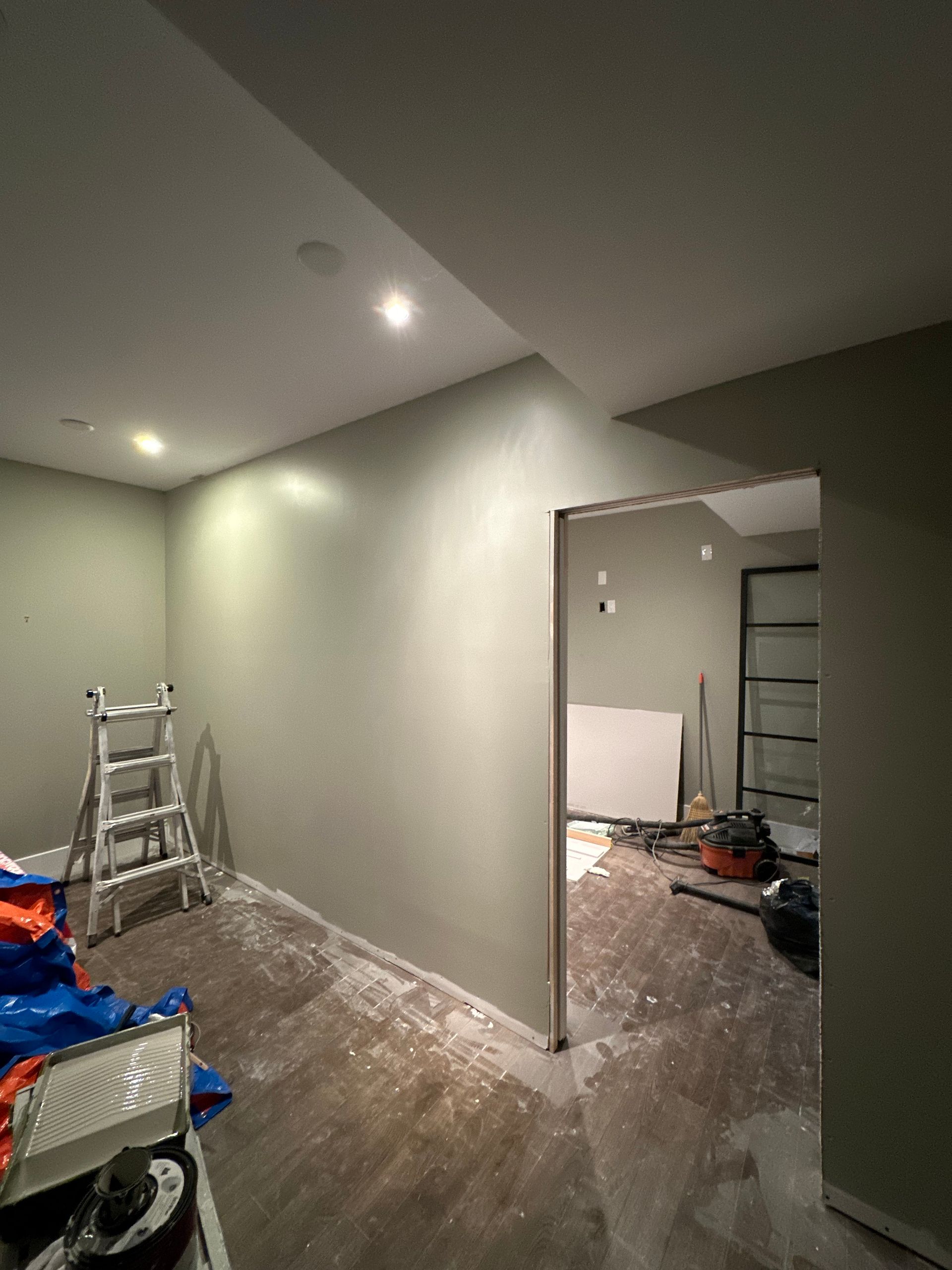 A remodel stopped at the finish stage: painted walls and ceiling with recessed lights on, a raw door jamb with no casing, no baseboard, outlet and switch boxes without plates, and a ladder, shop vacuum and tarps on a dusty plank floor