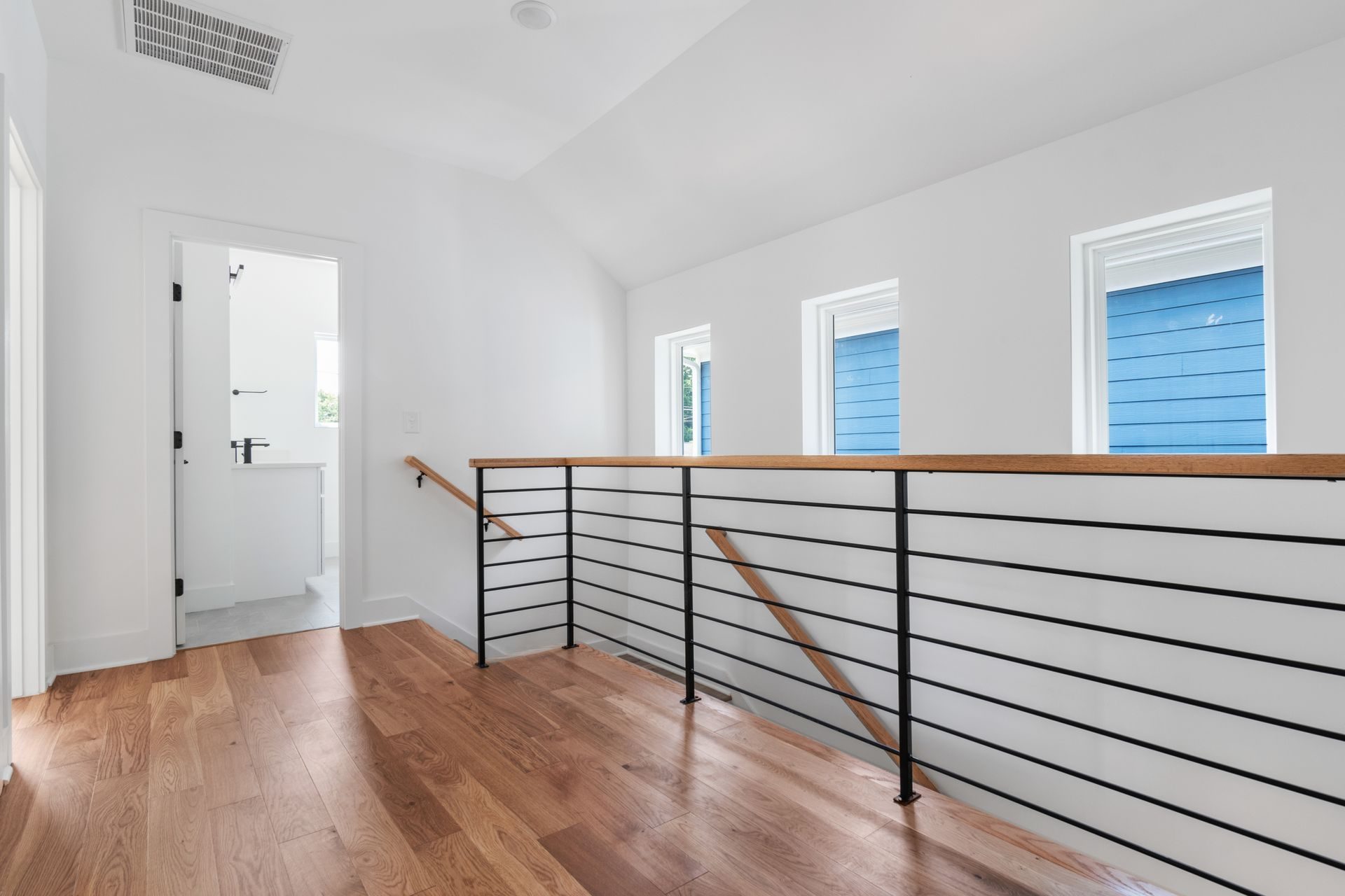 An empty upstairs landing at the top of a stair, with a sloped white ceiling, a wide-plank oak floor, a black horizontal-bar railing with a wood cap, three small windows on the outside wall and an open door to a white bathroom