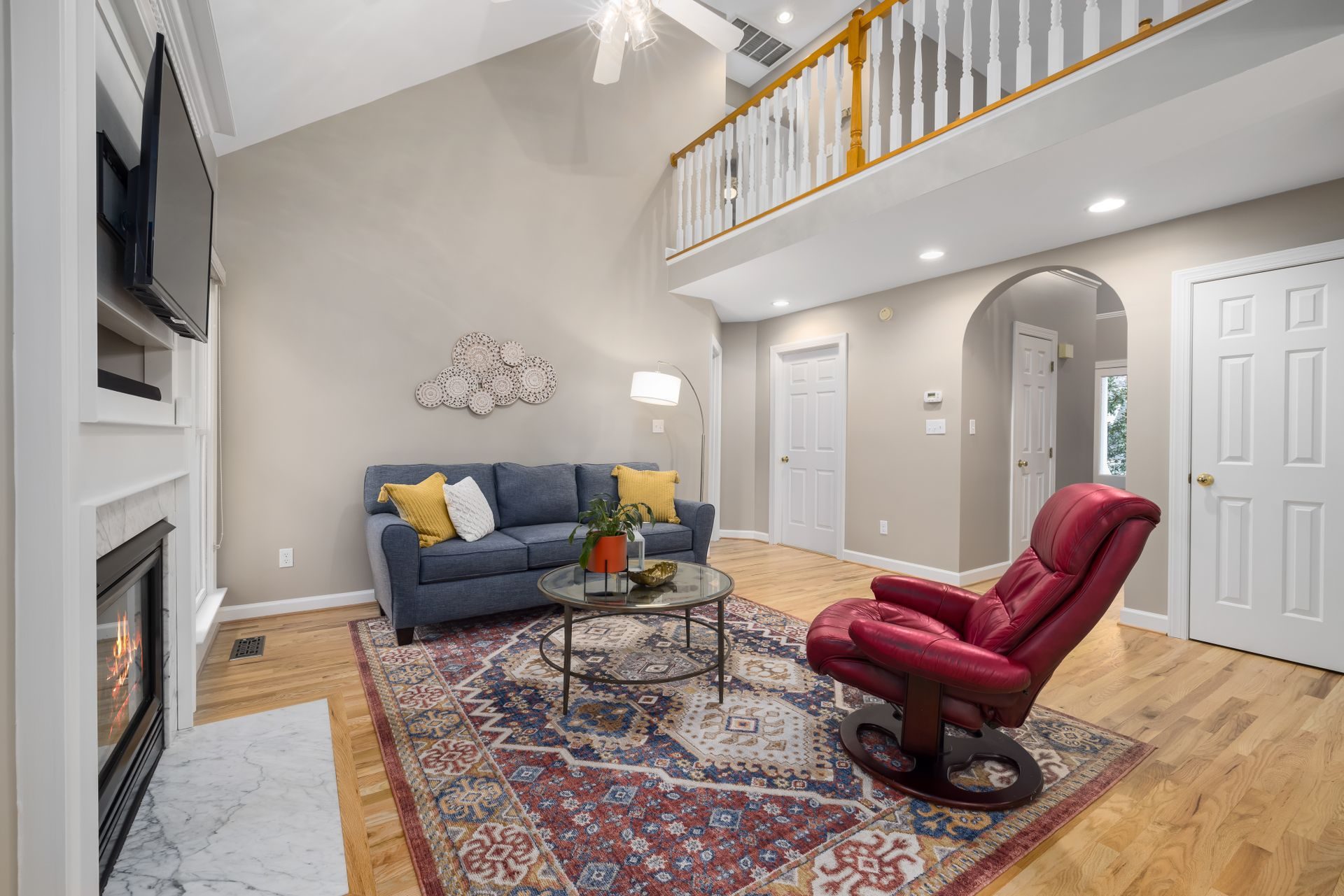 A vaulted living room with oak floors and a gas fireplace, a rounded drywall arch opening to a hall at the back, and an upstairs balcony railed with white balusters