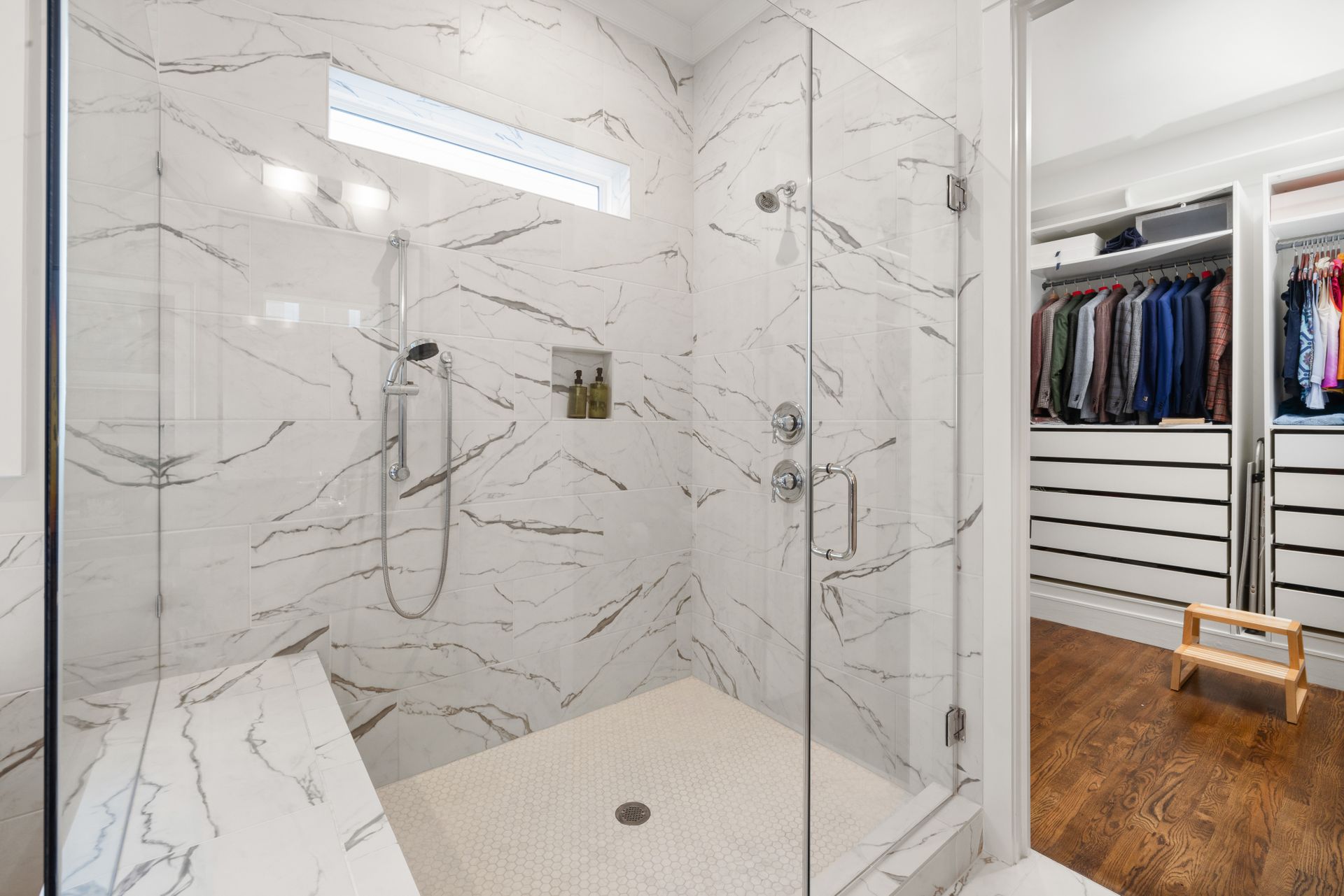Walk-in shower with a handheld shower on a polished-chrome slide bar beside a fixed shower head, two round valve trims, a long tiled bench, a recessed niche and a white hexagon mosaic floor