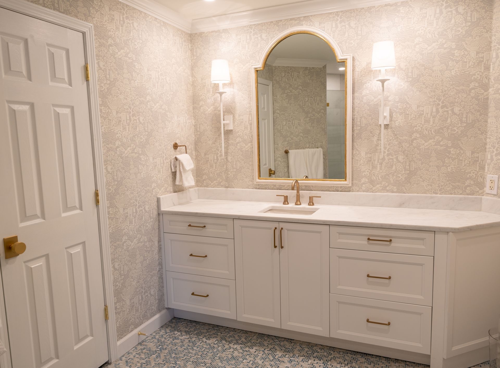 A long white shaker vanity with brass pulls, a white marble-look top and a single undermount sink with a brushed-brass widespread faucet, under an arched brass-framed mirror and two wall sconces