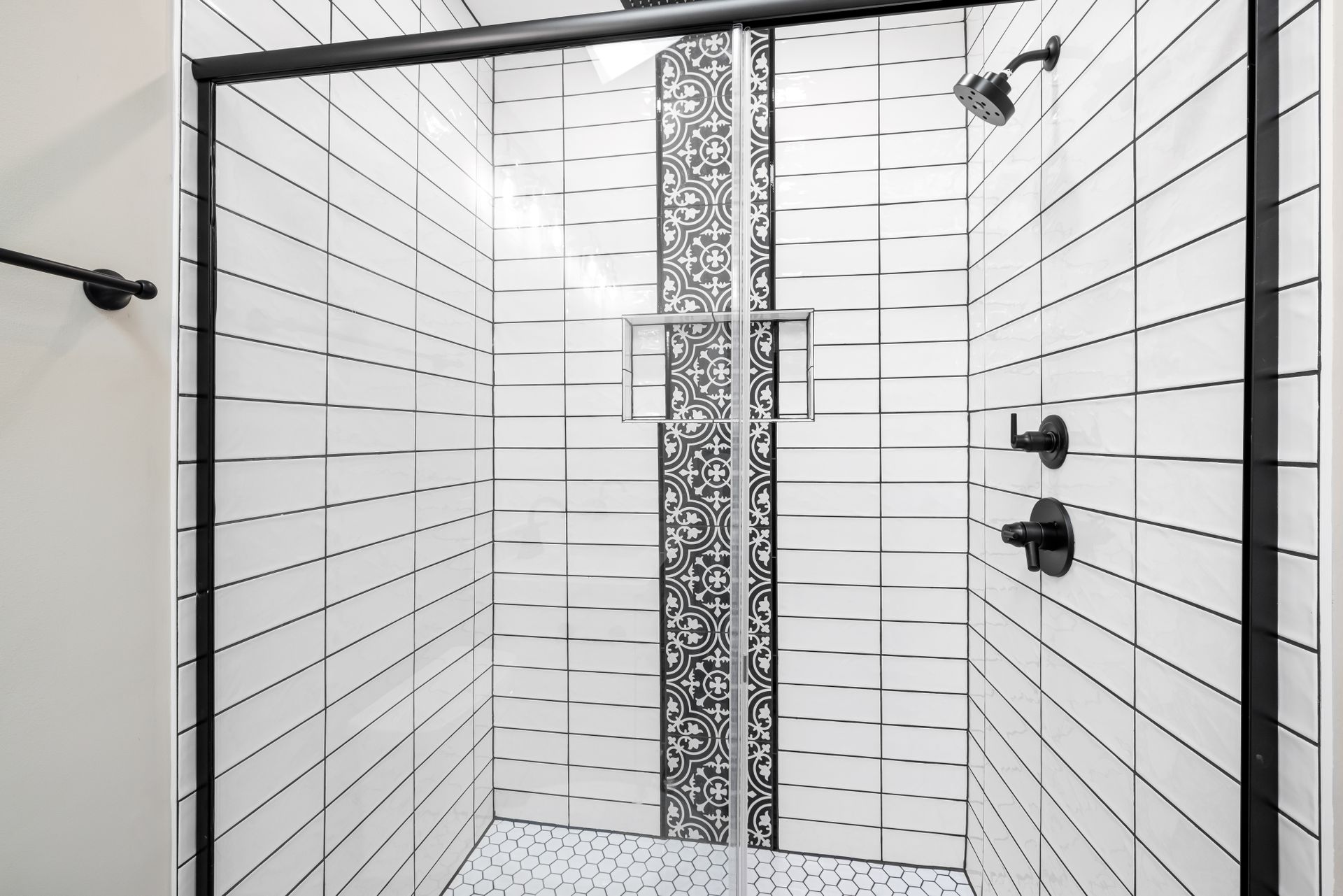 Shower alcove tiled floor to ceiling in glossy white rectangular tile with dark grout, a vertical band of black-and-white patterned tile running up through a recessed niche, a white hexagon mosaic floor, matte-black fixtures and a black-framed sliding glass door