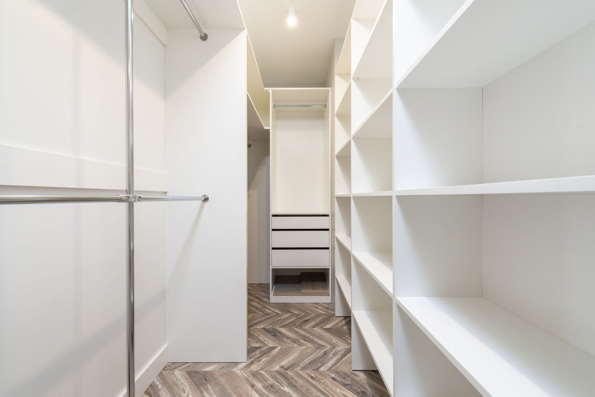 A white walk-in closet with modular open shelving on the right, chrome hanging rods on the left and a white drawer unit at the far end, no doors fitted, over a herringbone wood-look floor