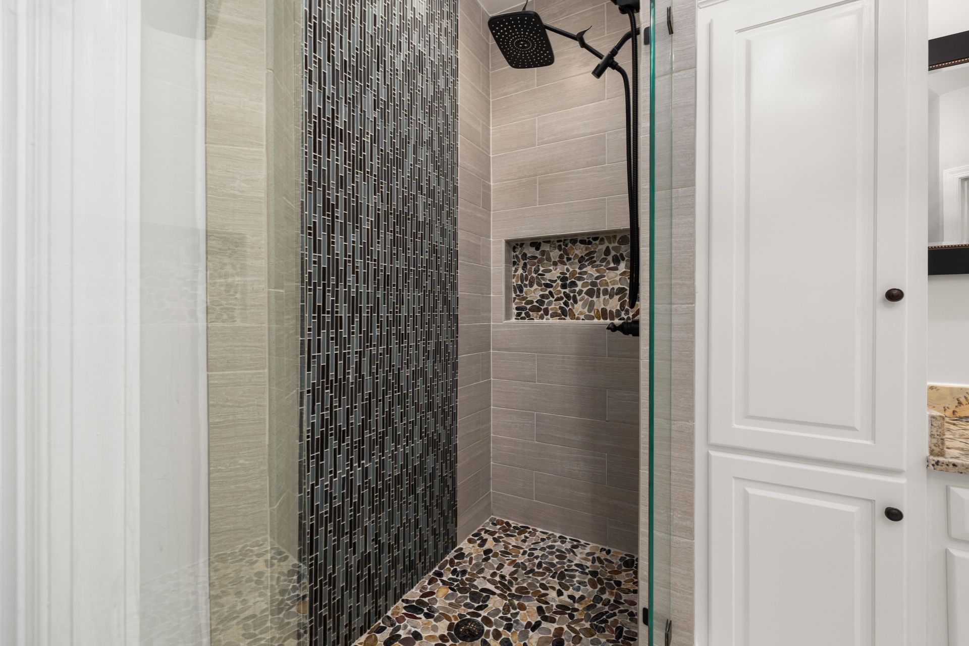 Shower with gray wood-grain porcelain plank walls, a full-height strip of dark gray and blue glass mosaic, a river-pebble mosaic floor and pebble-lined niche, a matte-black rain head and handheld, and a frameless glass door