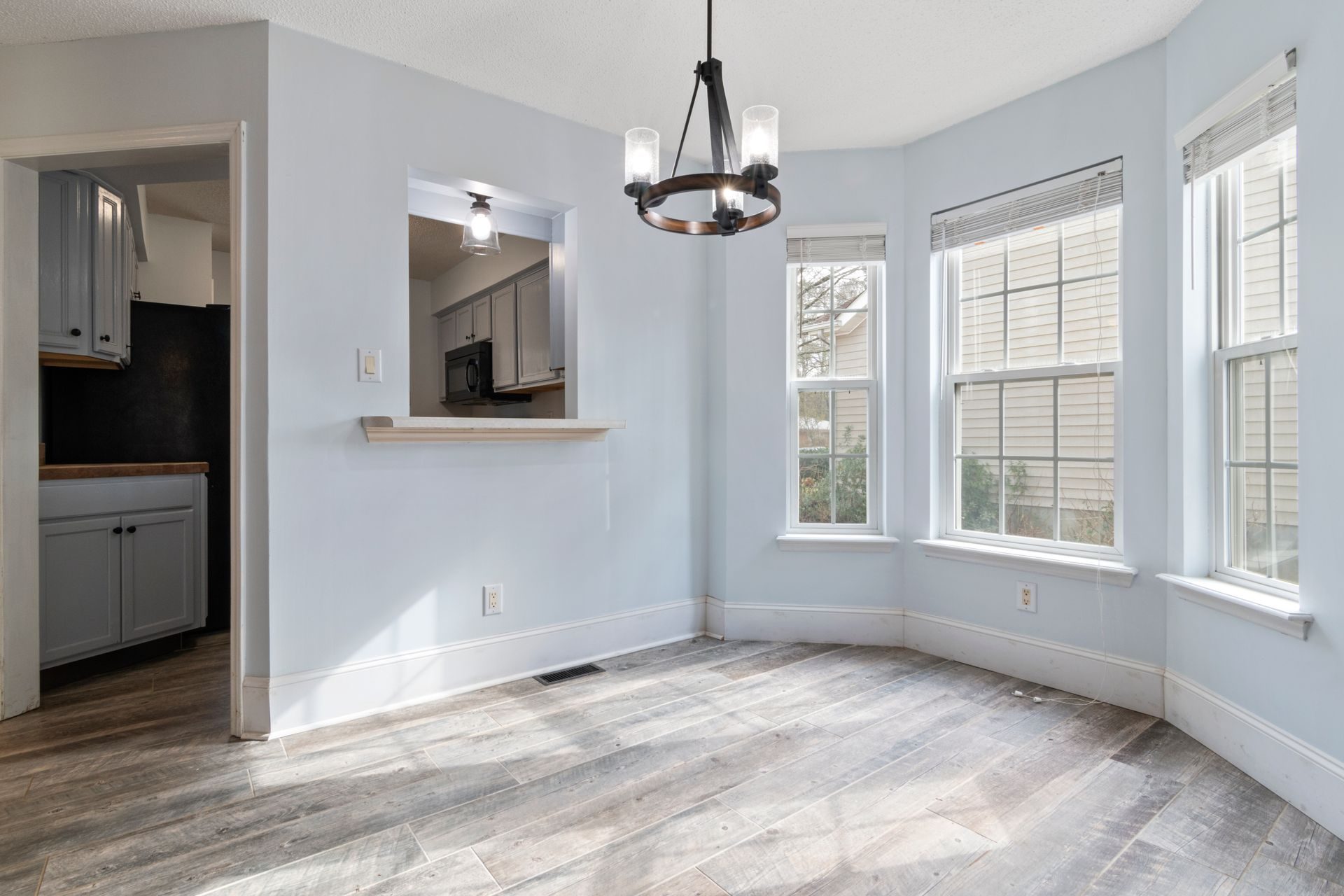 An empty, sunlit dining nook with a gray-brown wood-look plank floor laid lengthwise, a three-window bay and a black iron chandelier, open to a kitchen with gray cabinets