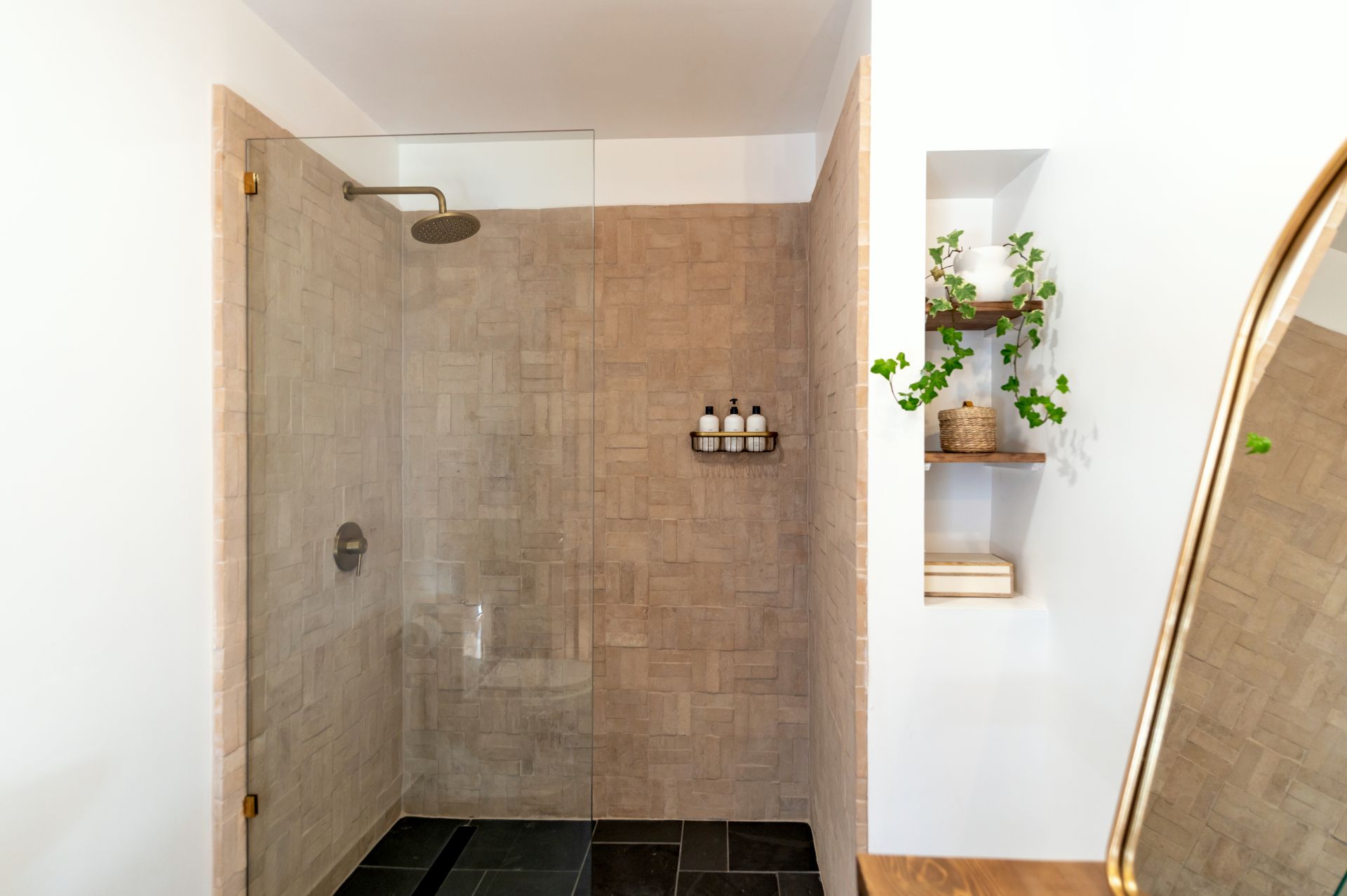 Walk-in shower lined in handmade blush-beige zellige-style tile laid in a basketweave, with a fixed frameless glass panel on brushed-brass clips, a brushed-brass rain head and a black slate floor with a linear drain